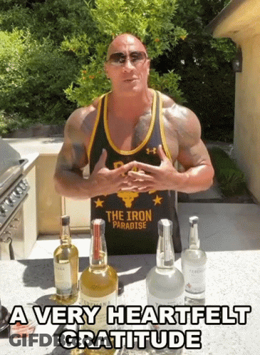 A Man Wearing A Tank Top That Says The Iron Paradise Stands In Front Of Bottles Of Alcohol GIF