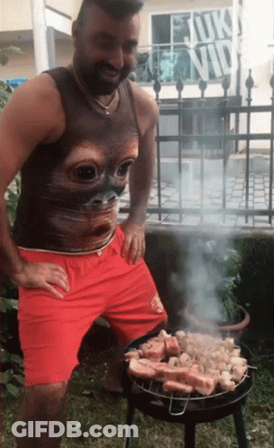 A Man Wearing A Tank Top With A Cat Face On It Is Cooking Meat On A Grill GIF