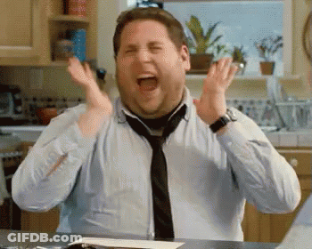 A Man Wearing A Tie And A Blue Shirt Is Sitting At A Table With His Hands In The Air . GIF