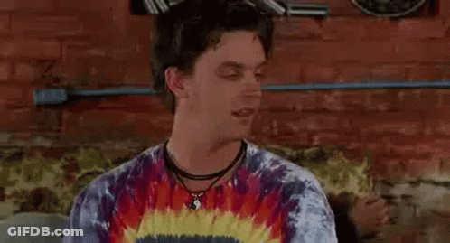 A Man Wearing A Tie Dye Shirt And A Choker Is Sitting On A Couch In Front Of A Brick Wall . GIF