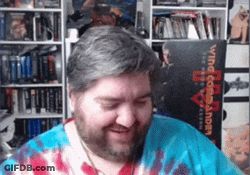 A Man Wearing A Tie Dye Shirt Is Smiling In Front Of A Book Titled Wing Commander GIF