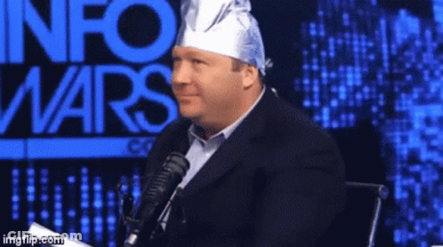 A Man Wearing A Tin Foil Hat Is Sitting In Front Of A Microphone In Front Of An Info Wars Sign GIF