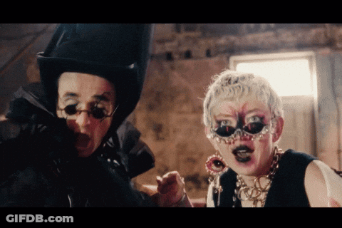 A Man Wearing A Top Hat And Glasses Stands Next To A Woman Wearing A Mask And Earrings GIF