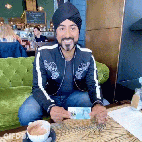 A Man Wearing A Turban Is Sitting At A Table Holding A Dollar Bill GIF