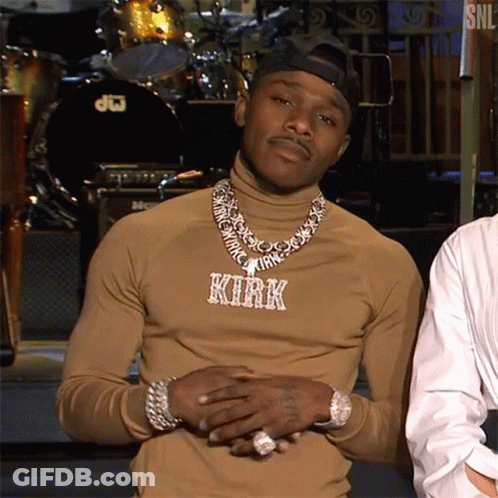 A Man Wearing A Turtleneck And A Necklace With The Name Kirk On It Is Standing In Front Of A Drum Set . GIF