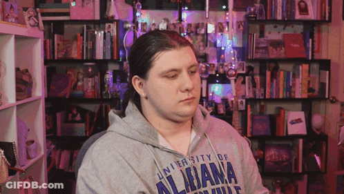 A Man Wearing A University Of Alabama Sweatshirt Sits In Front Of A Bookshelf GIF