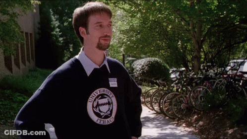 A Man Wearing A University Of Ithaca Sweater Stands On A Sidewalk GIF