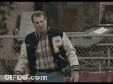 Man In Varsity Jacket Standing By Fence GIF