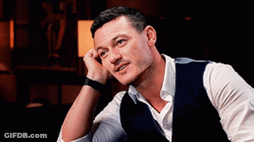 A Man Wearing A Vest And A White Shirt Is Sitting Down With His Hand On His Chin . GIF