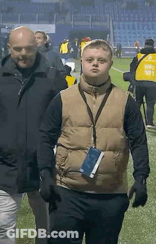 A Man Wearing A Vest With A Lanyard Around His Neck Is Walking On A Field . GIF