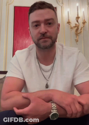 A Man Wearing A Watch And A Necklace Is Sitting With His Hands Folded GIF