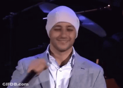 A Man Wearing A White Beanie And A Suit Is Smiling And Holding A Microphone GIF