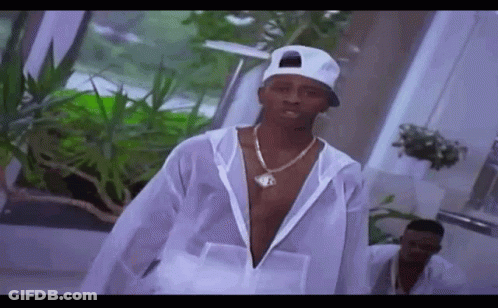 A Man Wearing A White Hat And A Necklace Is Standing In A Room With Plants . GIF
