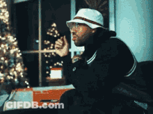 A Man Wearing A White Hat And Glasses Is Sitting In Front Of A Christmas Tree . GIF