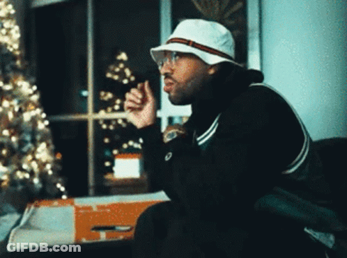 A Man Wearing A White Hat And Glasses Sitting In Front Of A Christmas Tree GIF