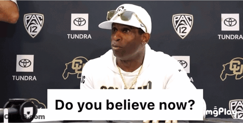 A Man Wearing A White Hat And Sunglasses Is Sitting In Front Of A Sign That Says " Do You Believe Now " GIF