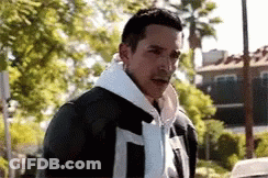 A Man Wearing A White Hoodie And A Black Leather Jacket Is Standing In Front Of A Palm Tree . GIF