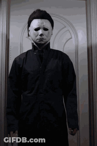 A Man Wearing A White Mask And A Black Jacket Is Standing In Front Of A Door GIF