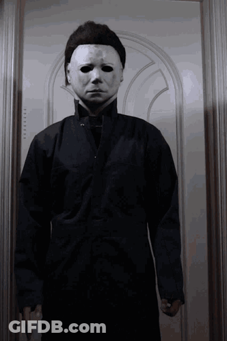 A Man Wearing A White Mask And A Black Jacket Is Standing In Front Of A Door GIF