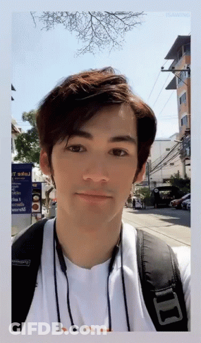 A Man Wearing A White Shirt And A Black Backpack Stands In Front Of A Sign That Says ' T ' On It GIF