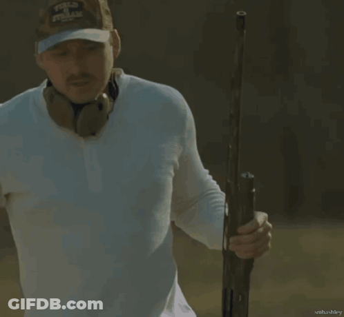 A Man Wearing A White Shirt And A Camo Hat Holds A Gun In His Right Hand GIF