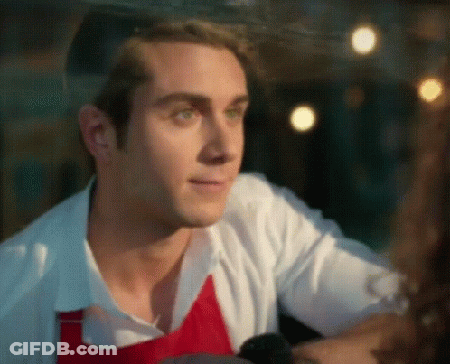 A Man Wearing A White Shirt And A Red Apron Is Looking Out A Window GIF