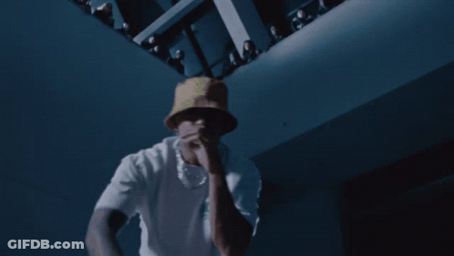 A Man Wearing A White Shirt And A Yellow Hat Is Dancing In A Dark Room GIF