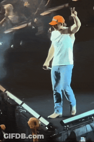 A Man Wearing A White Shirt And An Orange Hat Stands On A Stage Giving A Peace Sign GIF