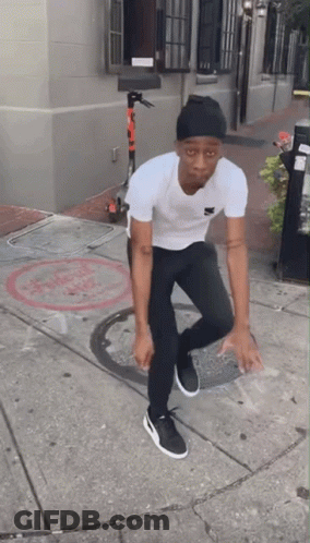 A Man Wearing A White Shirt And Black Pants Is Standing On A Sidewalk GIF