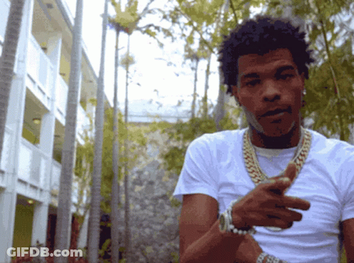 A Man Wearing A White Shirt And Gold Chains Is Standing In Front Of A Building GIF