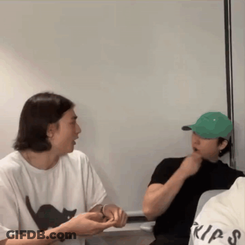 A Man Wearing A White Shirt With A Black Cat On It Is Talking To Another Man Wearing A Green Hat GIF