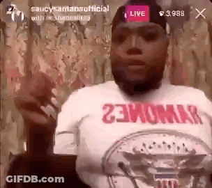 A Man Wearing A White Shirt With Red Writing On It Is Sitting In Front Of A Microphone And Talking . GIF