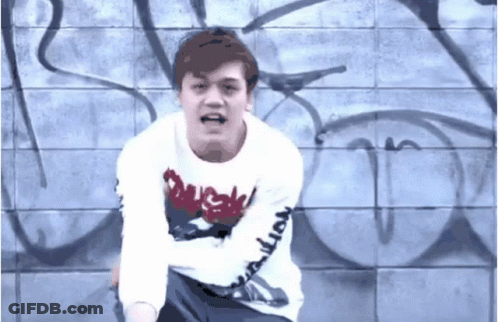 A Man Wearing A White Shirt With The Word Duck On It Stands In Front Of A Wall With Graffiti On It GIF