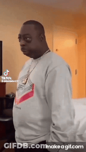 A Man Wearing A White Sweater And A Necklace Is Standing In A Room With His Hands In His Pockets . GIF