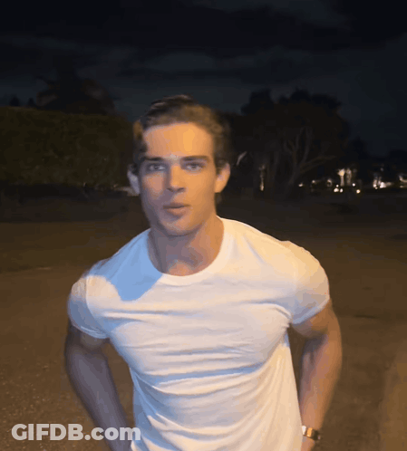 A Man Wearing A White T-Shirt And A Watch Stands In The Dark GIF