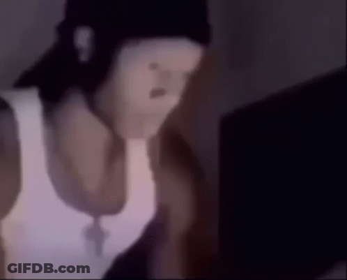 A Man Wearing A White Tank Top And A Black Hat Is Looking At A Laptop . GIF