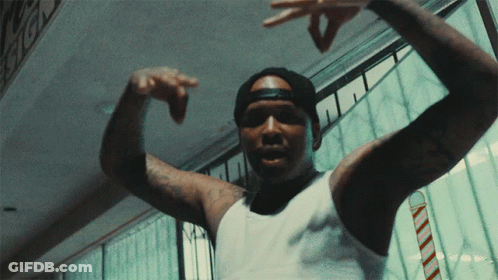A Man Wearing A White Tank Top And A Black Hat Is Pointing His Fingers GIF