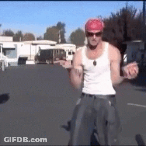 A Man Wearing A White Tank Top And A Red Hat Is Standing On A Bicycle . GIF