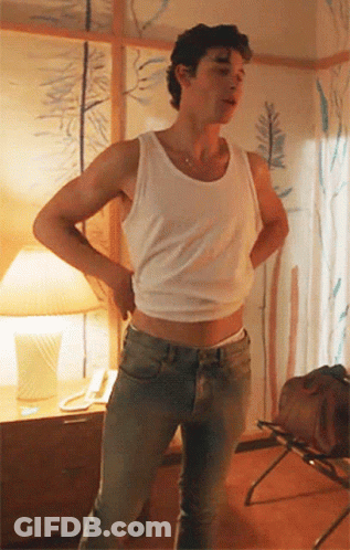A Man Wearing A White Tank Top And Jeans Is Standing In A Room With His Hands On His Hips . GIF