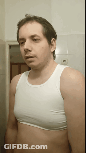 A Man Wearing A White Tank Top Stands In Front Of A White Tiled Wall GIF