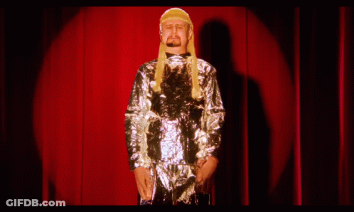 A Man Wearing A Wig And A Silver Jacket Stands On A Stage In Front Of A Red Curtain GIF
