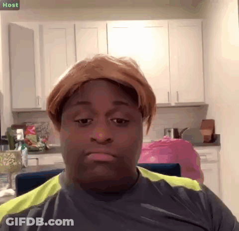 A Man Wearing A Wig Is Sitting In A Kitchen With A Sign That Says Host Above Him GIF