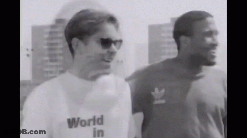 A Man Wearing A World In T-Shirt Is Standing Next To Another Man Wearing Sunglasses . GIF