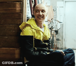 A Man Wearing A Yellow And Black Jacket Is Smiling While Leaning Against A Wooden Wall GIF