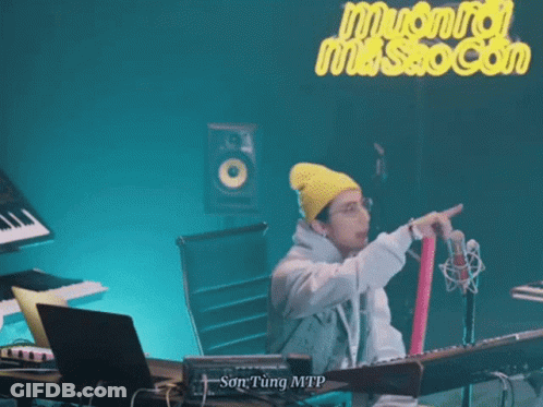 A Man Wearing A Yellow Beanie And Glasses Is Standing In Front Of A Microphone . GIF