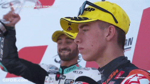 A Man Wearing A Yellow Hat With Leopards On It Stands Next To Another Man GIF