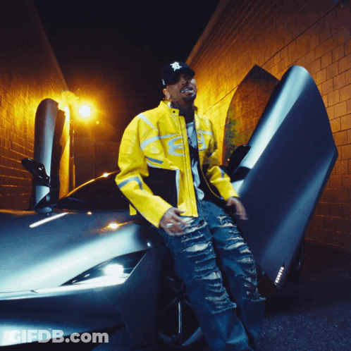 A Man Wearing A Yellow Jacket And A Black Hat Stands Next To A Car GIF