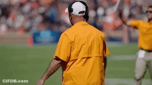 A Man Wearing A Yellow Jacket And A White Hat Stands On A Football Field GIF