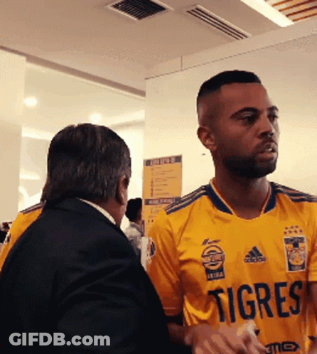 A Man Wearing A Yellow Jersey That Says Tigres Talks To Another Man GIF