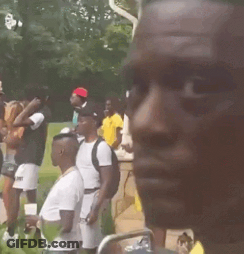A Man Wearing A Yellow Shirt And A Gold Chain Is Standing In Front Of A Crowd Of People . GIF